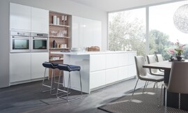 La pared de armarios empotrados con tiradores incrustados donde una estantería rompe la continuidad de los armarios de cocina y crea en esta cocina blanca con isla el ambiente de sala de estar. Zuordnung: Stil Cocinas de diseño, Planungsart Cocinas con isla