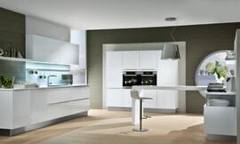 Esta cocina está pensada para ser el centro de una sala de estar. Una pared de armarios columna preside la sala. En ella están integrados los electrodomésticos. Una isla en el centro de la sala, que destaca por su sencillez y que a primera vista nos hace pensar que se trata de una mesa, es la superficie de trabajo principal con una amplia encimera de cristal y la placa de cocción integrada. A un lado, los armarios altos con el fregadero nos recuerdan a las cocinas tradicionales. Al otro, una cómoda donde descansa la isla nos acerca al ambiente de sala de estar. Los frentes blanco polar lacados de alto brillo le otorgan al conjunto el brillo de una cocina de gran calidad. Zuordnung: Stil Cocinas modernas, Planungsart Cocinas con office