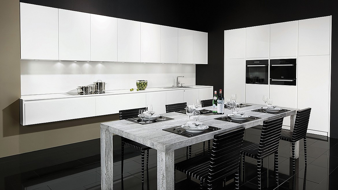 Cocina blanca de puertas sin tiradores de elegantes líneas de diseño. Los tiradores han sido sustituidos por elegantes acanaladuras. Junto a la perfección del color blanco destaca la encimera de Glasline, de vidrio esmaltado de color negro. Los paneles correderos redondean la perfección de cajones y puertas de esta cocina con isla. Zuordnung: Stil Cocinas de diseño, Planungsart Cocinas en L