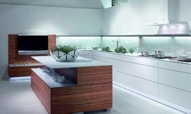 Cocina abierta de líneas puristas de Warendorf. El contraste de esta cocina en blanco puro y madera de tineo clara ayudan a asimilar dos conceptos y dos ambientes, uniendo la cocina con la sala de estar. La zona de trabajo en blanco puro, con una iluminación elegante y funcional, aparece delimitada de la zona de transición, delimitada por los frentes de madera de tineo. Zuordnung: Stil Cocinas de diseño, Planungsart Cocinas con isla