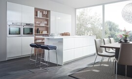 La pared de armarios empotrados con tiradores incrustados donde una estantería rompe la continuidad de los armarios de cocina y crea en esta cocina blanca con isla el ambiente de sala de estar. Zuordnung: Stil Cocinas de diseño, Planungsart Cocinas en L