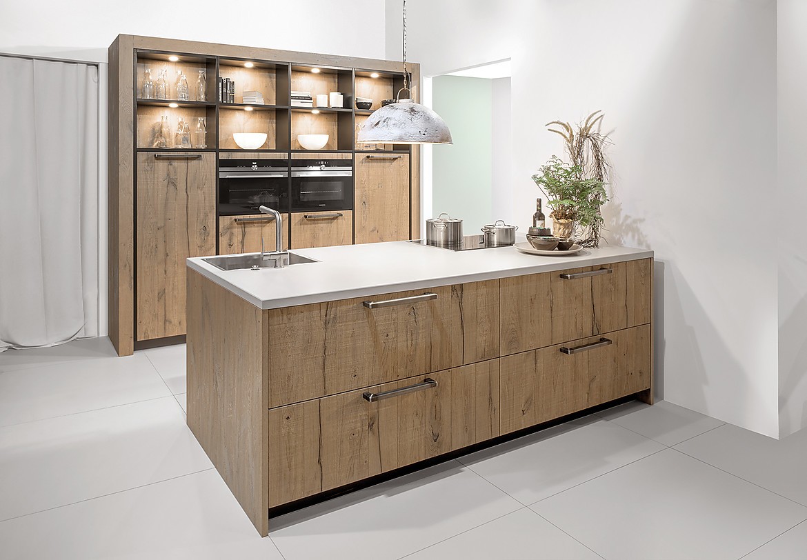 Esta cocina con isla con el frente de madera decorativa con óptica en Pastel Smoke hace realidad sus sueños. Los frentes de la cocina con una veta claramente visible, lo que le da un encanto un poco rústico a la cocina. Por encima de los electrodoésticos la cocina se ha complementado con una atractiva estantería iluminada. Zuordnung: Stil Cocinas de diseño, Planungsart Cocinas con isla