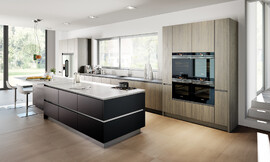 Esta cocina de diseño ofrece suficiente superficie de trabajo. Tiene una gran isla en negro mate con barra de desayuno y asientos que acaba con una encimera blanca. En el diseño de la zona del lavabo se recurrió a un frente de madera clara. Zuordnung: Stil Cocinas de diseño, Planungsart Cocinas en línea
