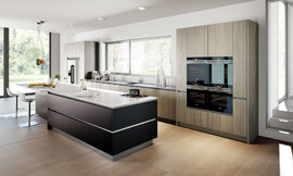 Esta cocina de diseño ofrece suficiente superficie de trabajo. Tiene una gran isla en negro mate con barra de desayuno y asientos que acaba con una encimera blanca. En el diseño de la zona del lavabo se recurrió a un frente de madera clara. Zuordnung: Stil Cocinas modernas, Planungsart Detalles del diseño