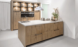 Esta cocina con isla con el frente de madera decorativa con óptica en Pastel Smoke hace realidad sus sueños. Los frentes de la cocina con una veta claramente visible, lo que le da un encanto un poco rústico a la cocina. Por encima de los electrodoésticos la cocina se ha complementado con una atractiva estantería iluminada. Zuordnung: Stil Cocinas de diseño, Planungsart Cocinas con office